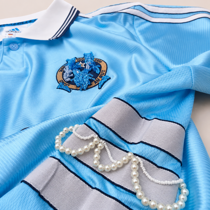 Détail d’un maillot de foot bleu ciel upcyclé OHBAB avec volants argentés, cascades de perles blanches et écusson brodé de perles bleues.