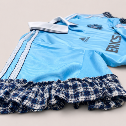 Détail d’un maillot de foot bleu ciel upcyclé OHBAB avec volants argentés, cascades de perles blanches et écusson brodé de perles bleues.