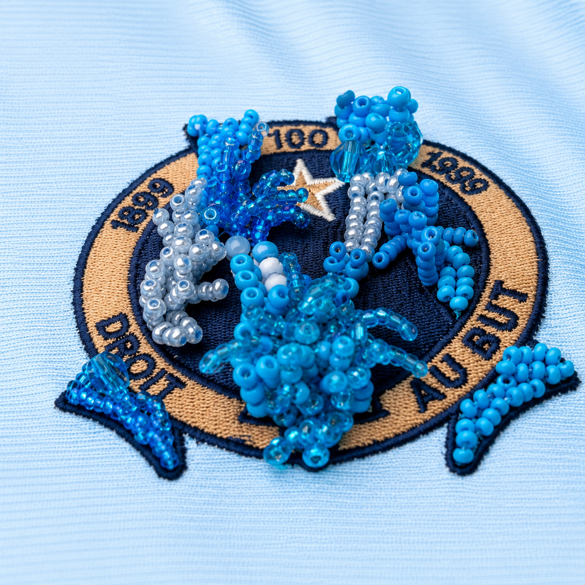 Détail d’un maillot de foot bleu ciel upcyclé OHBAB avec volants argentés, cascades de perles blanches et écusson brodé de perles bleues.